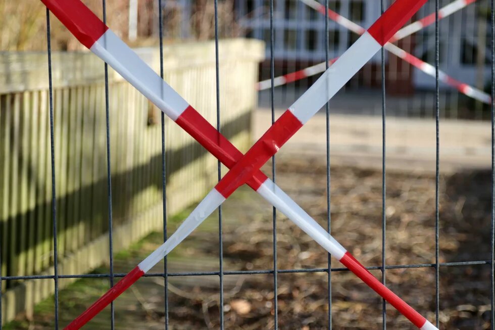 Foto: Ein Metallzaun mit rot-weißem Absperrband, das sich kreuzt. Dahinter ein Weg und ein Holzzaun, im Hintergrund unscharf ein Gebäude.
